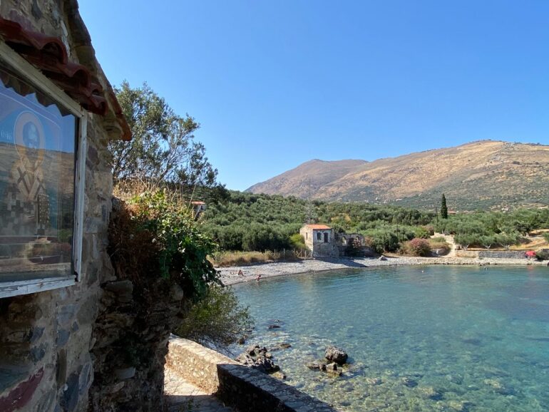 Ακρογιαλιά στην Καρδαμύλη με καταγάλανα νερά, πέτρινα σπίτια, ελαιώνες και βουνό στο βάθος